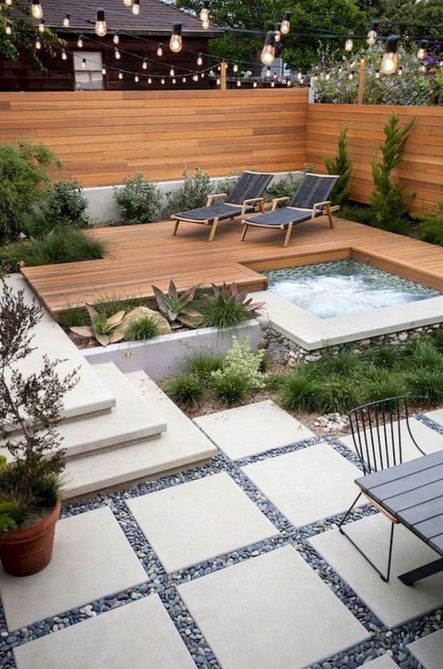 a small modern backyard with a stained fence and deck, with a hot tub, growing plants and loungers, with a dinign table and chairs
