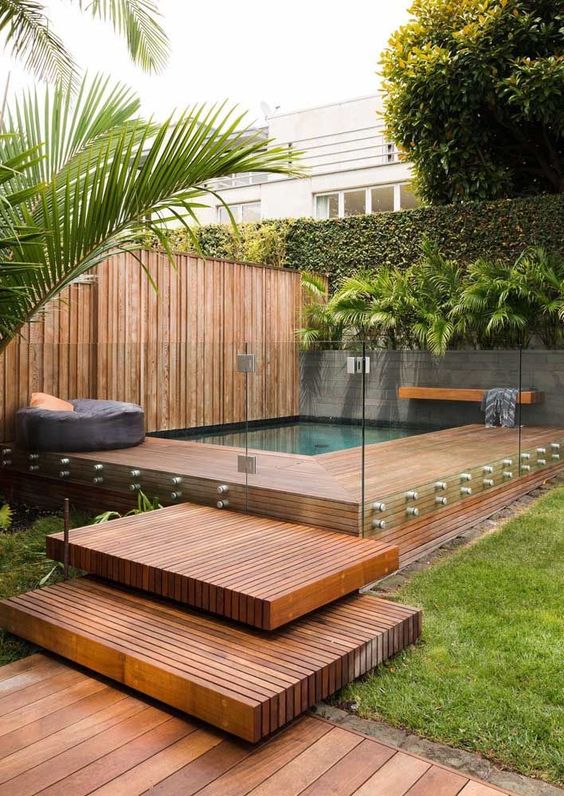 A tropical outdoor space with greenery, a rich stained deck and steps and a small pool with a bench and a pouf