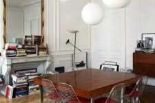 a Parisian dining room with a fireplace filled with books, a large mirror in a gilded frame, a stained storage unit, a stained table and Eames wire chairs plus pendant bubble lamps