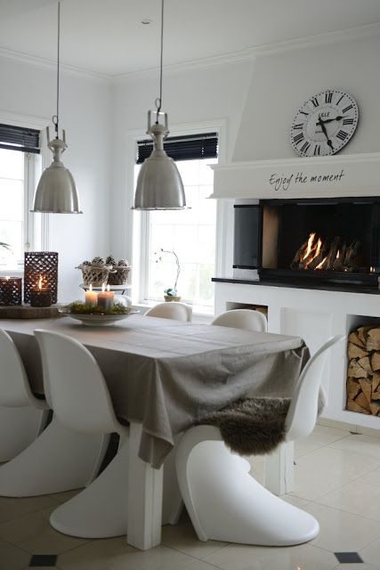 A Scandinavian dining room with a built in fireplace and firewood storage, a table and white Panton chairs, metal pendant lamps