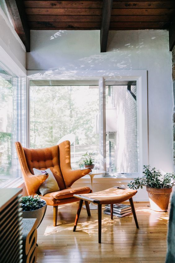 A beautiful rust colored Papa Bear chair with a pillow, a footrest, potted plants creates a cozy mid century modern nook