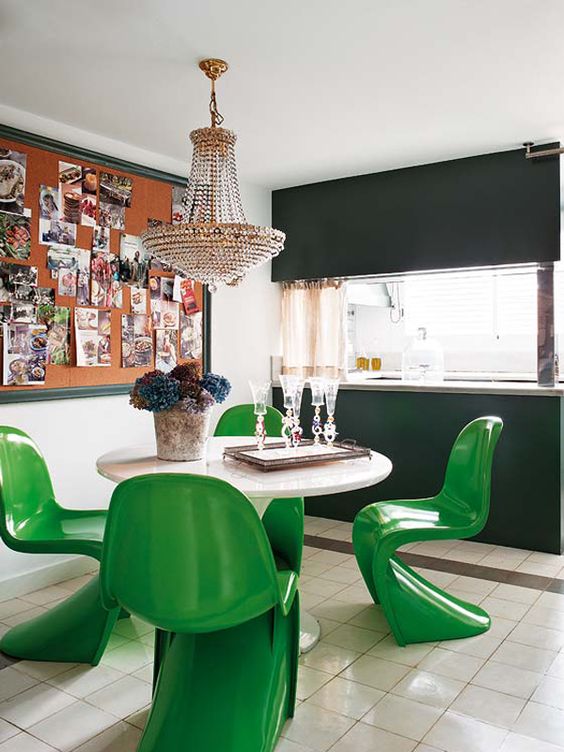 a bold dining space with a round table, bold green Panton chairs, a memo board with various photos and a crystal chandelier