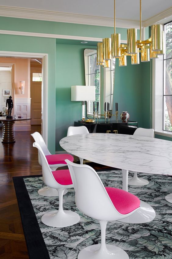 a bright and sophisticated dining room with green walls, an oval table, hot pink Tulip chairs, a gold chandelier and a glam credenza