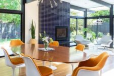 a bright mid-century modern dining room with an oval table and rust-colored tulips chairs, a fireplace clad with navy skinny tiles and a sunburst chandelier