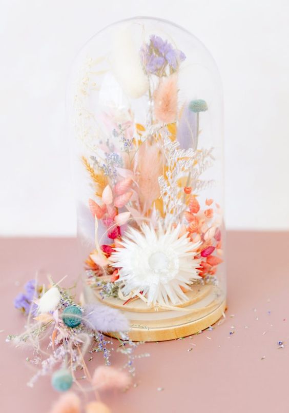 a bright pastel dried flower arrangement in pink, orange, lilac, red and neutral blooms and grasses in a neutral cloche