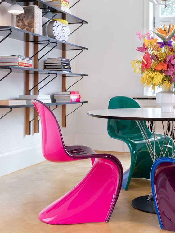 A catchy dining room with wall mounted shelves, a neutral chair and jewel tone Panton chairs plus bold bloos