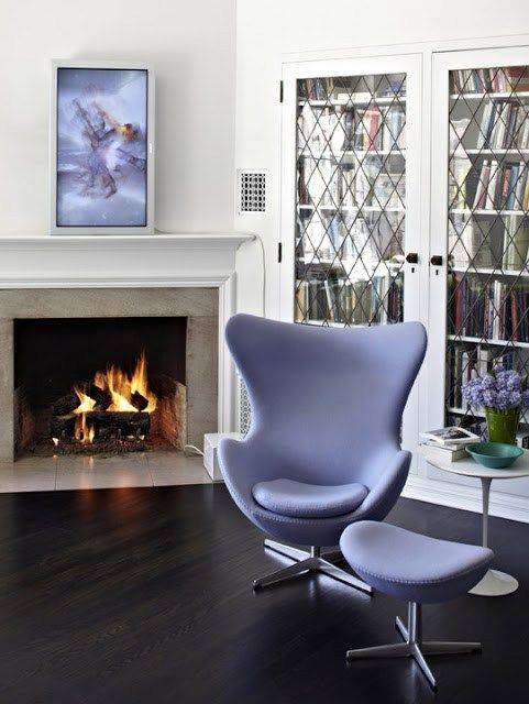 A chic space with a built in bookcase, a fireplace, a periwinkle Egg chair with a footrest, a side table and an artwork