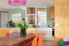a colorful dining room with a stained table, orange Panton chairs, a pink pendant lamp and some bright decor and blooms