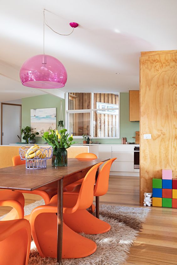 a colorful dining room with a stained table, orange Panton chairs, a pink pendant lamp and some bright decor and blooms