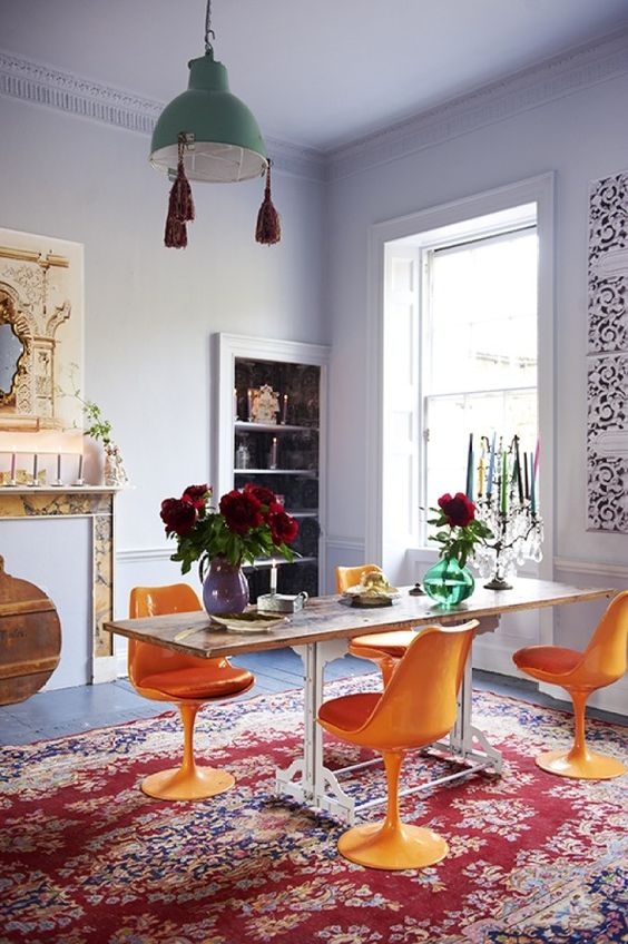 A colorful dining room with lilac walls and a ceiling, a fireplace, a built in storage unit, a table, orange Tulip chairs, a green pendant lamp