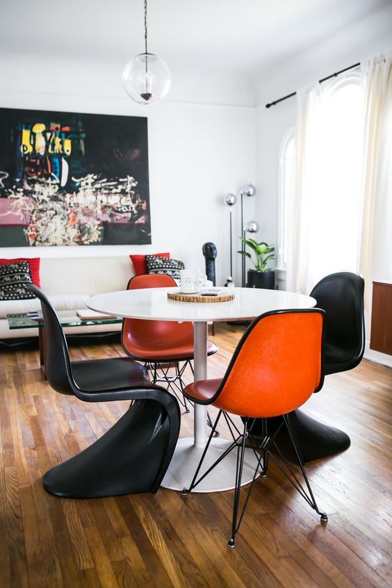 a contrasting dining room with a neutral sofa and bold pillows, an oversized artwork, a round table with Eames and Panton chairs in orange and black