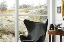 a cool space with a glazed wall, white beadboard walls, a dark-stained side table with decor and a black Egg chair plus some art