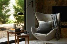 a cozy nook with a fireplace clad with stone, a grey Egg chair with a pillow, a couple of side tables and a gorgeous view