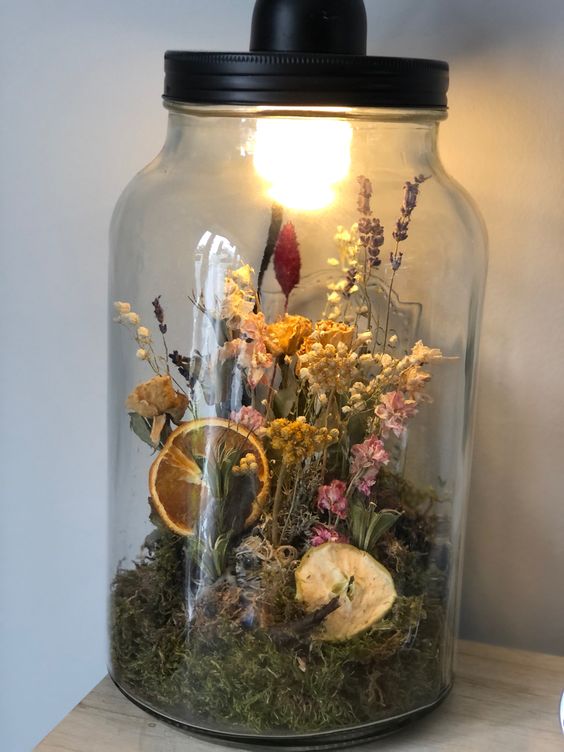 A jar with a black lid and a built in bulb, with moss, dried blooms, grasses and fruit slices, feels and looks like summer for real