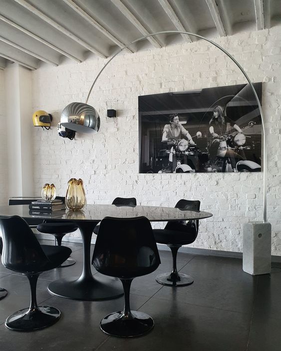A jaw dropping dining space with a white brick wall, an oval black table, black Tulip chairs, a floor lamp and a large photo