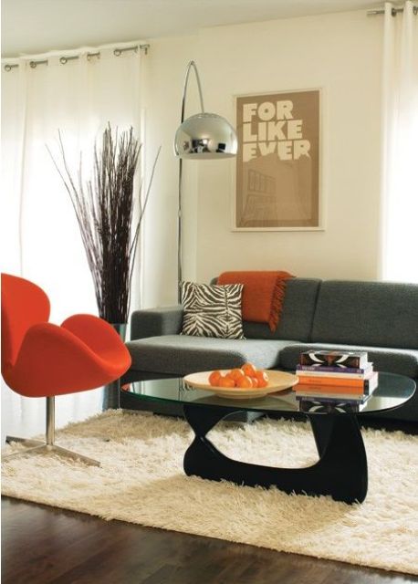 A mid century modern living room with a grey sofa and printed pillows, an orange Swan chair, a glass table and a neutral rug