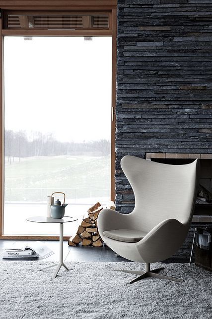 A neutral Egg chair, a white side table, a built in fireplace, a stack of firewood and a gorgeous view compose a lovely look