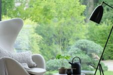 a peaceful nook with a glazed wall and a greenery view, a white Egg chair with pillows, a black pouf, a black floor lamp and a side table