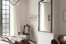 a refined Parisian bedroom with a bed and neutral bedding, a refined white chair and a brown leather Swan sofa, a mirror and a crystal chandelier