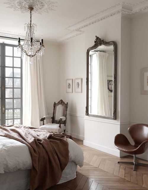 a refined Parisian bedroom with a bed and neutral bedding, a refined white chair and a brown leather Swan sofa, a mirror and a crystal chandelier