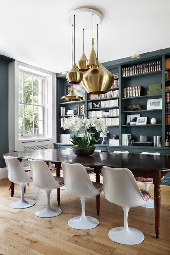 A sophisticated dining room with slate grey walls, a large built in storage unit, a black dining table and Tulip chairs plus a cluster of brass pendant lamps