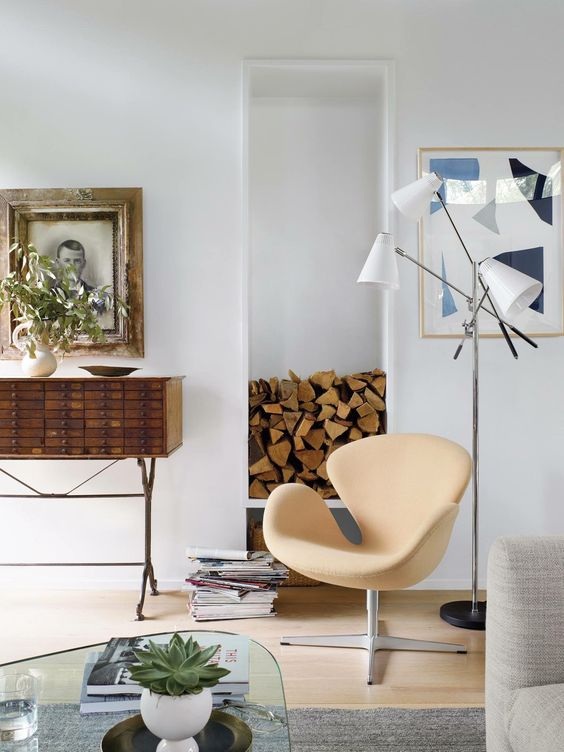 an airy space with a tan Swan chair, a firewood niche, a stained storage unit, an artwork and a floor lamp is cool
