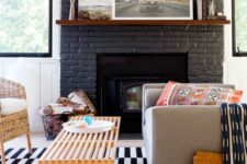 06 a black painted brick fireplace, a grey sofa, a woven chair, a wooden bench as a coffee table and some printed textiles