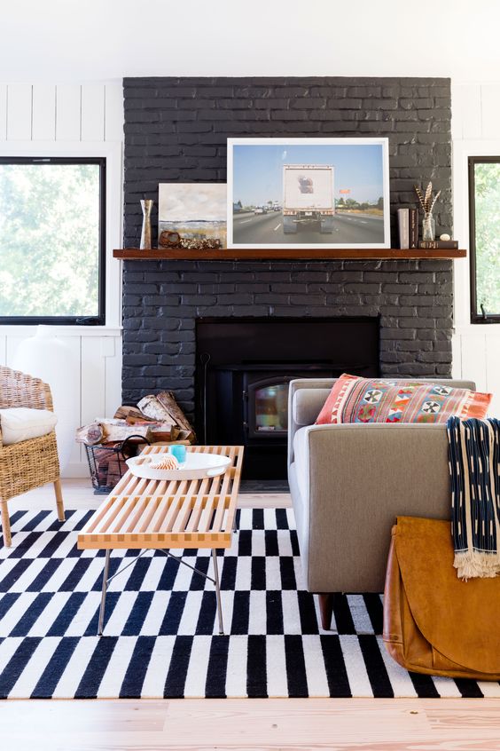 a black painted brick fireplace, a grey sofa, a woven chair, a wooden bench as a coffee table and some printed textiles