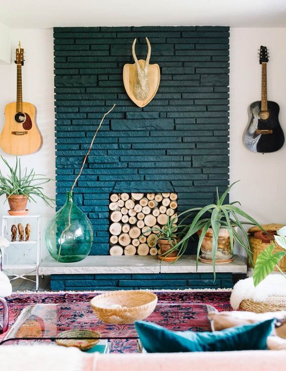 a bold boho living room with a navy painted brick fireplace, a wood log screen, potted plants, some decor, a bold rug and an acrylic table