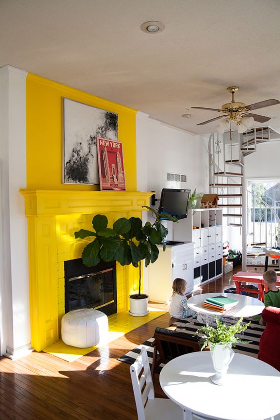 a modern living room with a bold yellow fireplace, a striped rug, some potted plants and some artwork