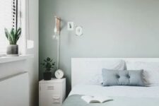 a Scandinavian bedroom with a white bed and green and white bedding, white metal cabinets, a pendant lamp and sconces