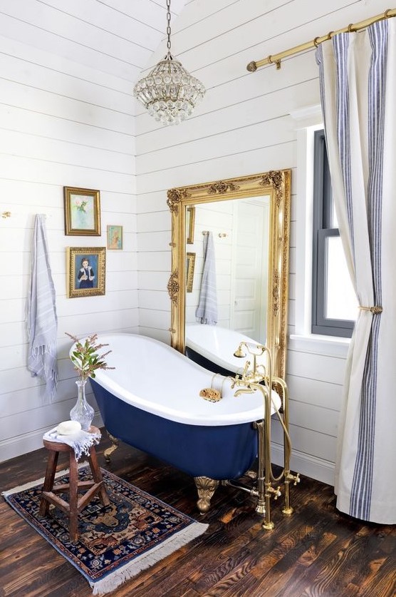 a bathroom covered with white shiplap, with a mirror in an ornate gold frame, artworks, blue tub on gold legs