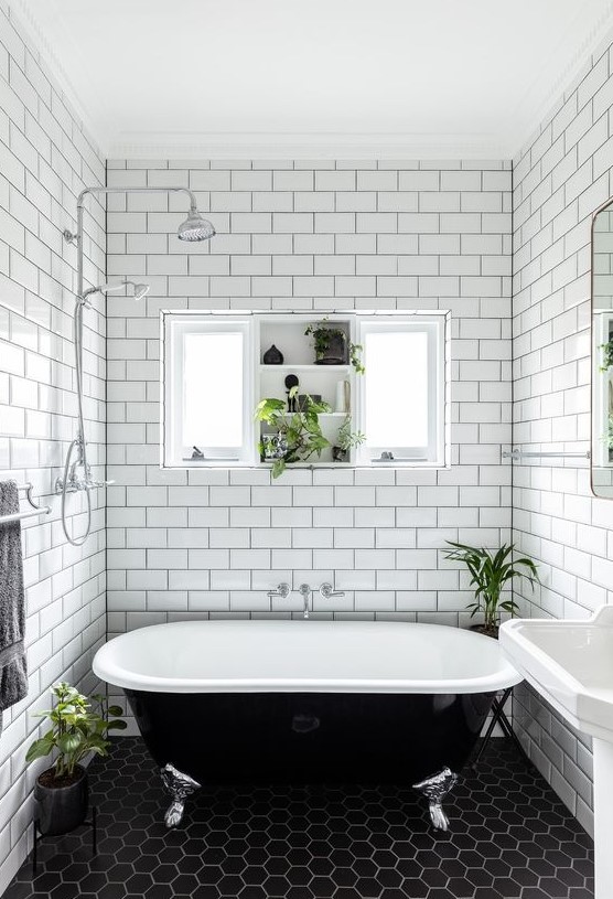 A beautiful black and white bathroom with white subway and black hex tiles, a free standing sink, a black vintage bathtub and a window