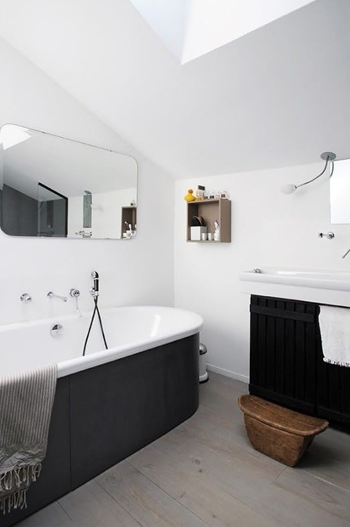 a contemporary black and white bathroom with white walls and laminate on the floor, a tub clad with black panels, a black paneled floating vanity and a rounded mirror