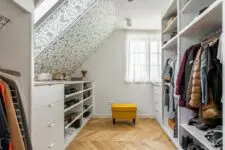 a cool attic closet with wallpaper walls and ceilings, dressers, open storage compartments and a yellow stool is a lovely space