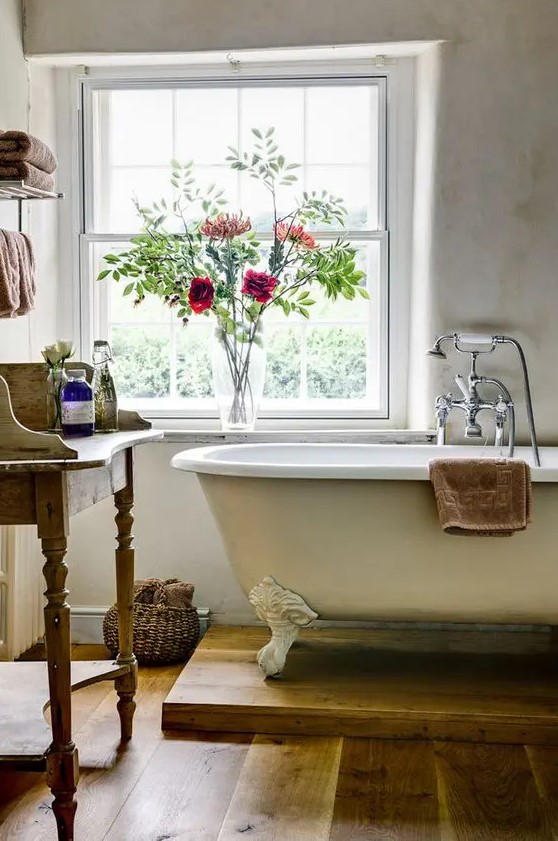 a country bathroom with neutral walls, a neutral clawfoot bathtub, a wooden vanity and blooms and towels