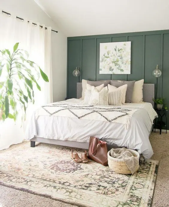 a cozy farmhouse bedroom with a green accent wall, a grey bed with neutral bedding, a printed rug, potted greenery and an artwork