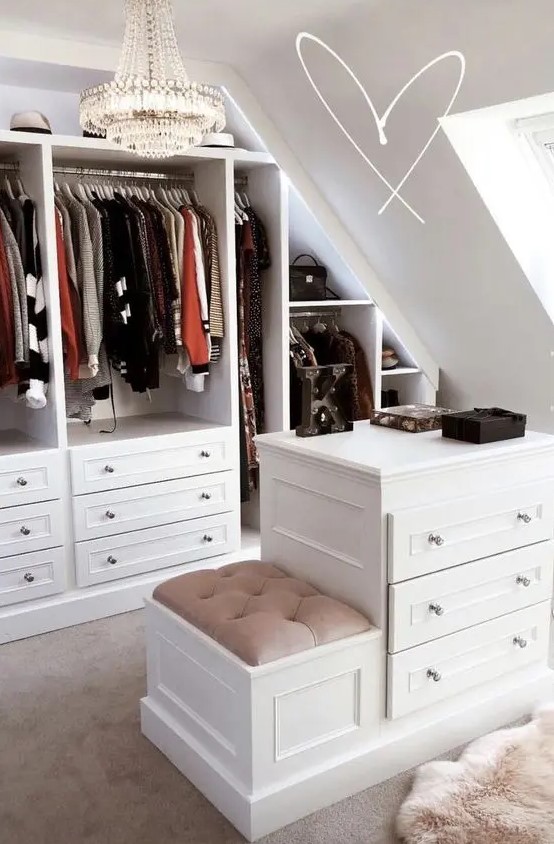 a glam sloped closet with open shelves and drawers, a dresser with a stool, a chic crystal chandelier is amazing