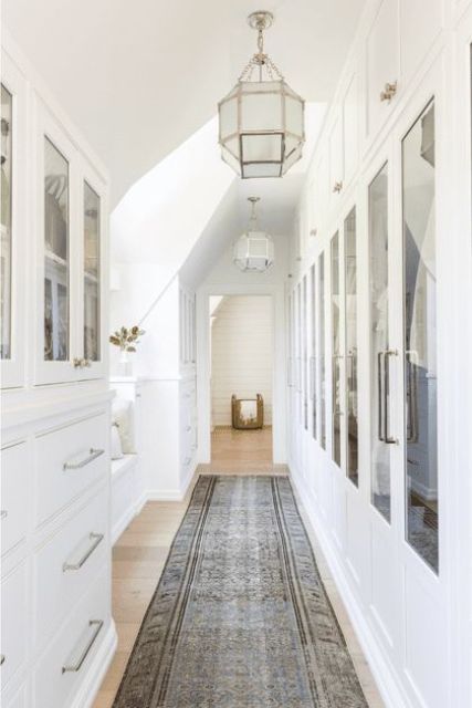 A large white farmhouse closet with built in wardrobes with glass doors, drawers, cabinets and a bench at the window is amazing
