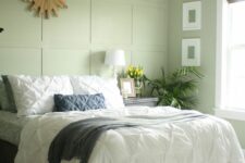 a light green bedroom with a paneled wall, a bed with neutral bedding, a nightstand, potted greenery, a burst mirror