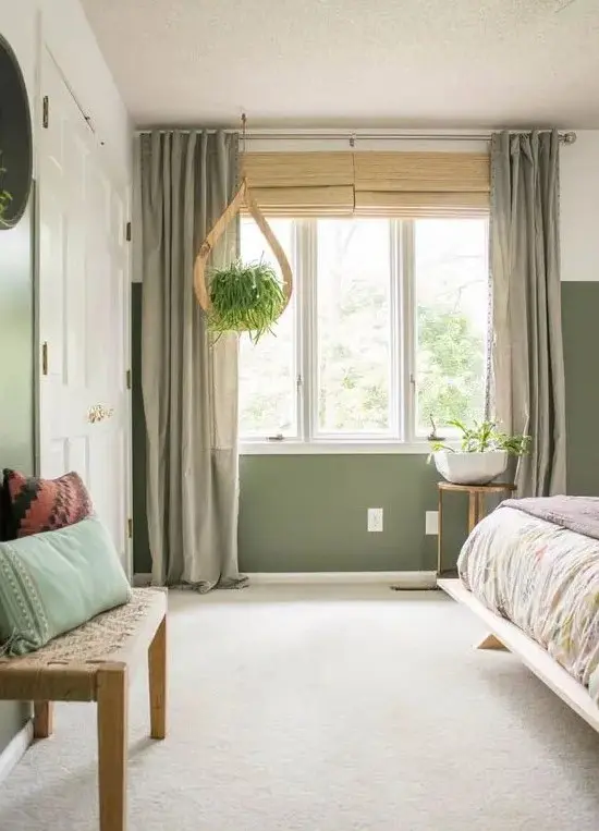 a lovely bedroom with green walls, a bed with printed bedding, a neutral floor and grey curtains, a woven bench and some plants