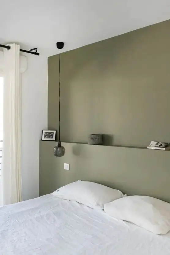a minimal bedroom with an olive green accent wall, a white bed and bedding, a pendant lamp and some decor