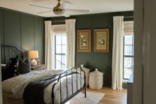 a moody vintage bedroom with dark green panaled walls, a black forged bed with neutral bedding, baskets and white curtains