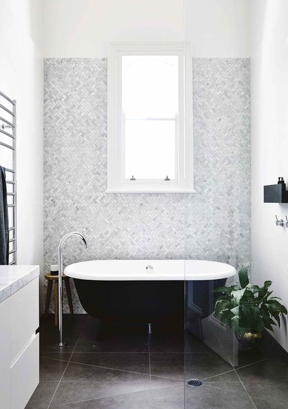 a neutral and chic bathroom with a grey marble accent wall, dark tiles on the floor, a black clawfoot tub, a white vanity and a black mini shelf
