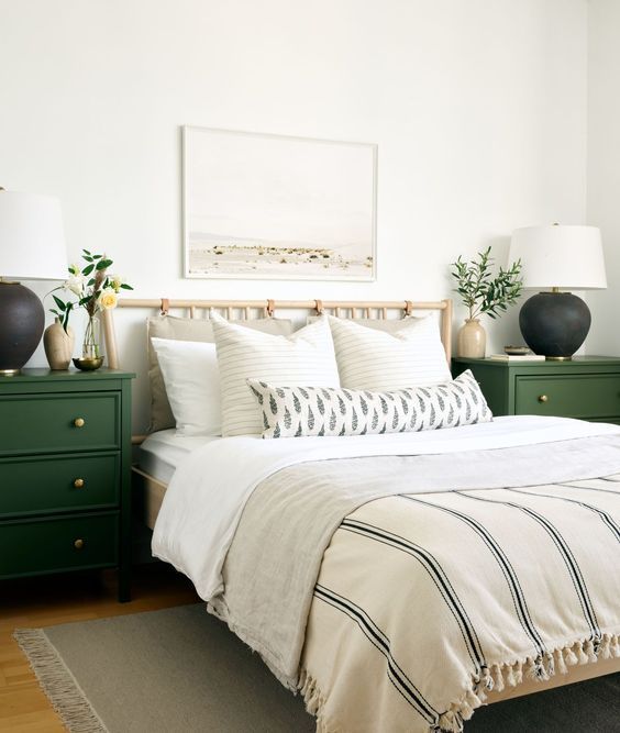 a neutral farmhouse bedroom with a wooden bed with neutral bedding, green nightstands, two-tone lamps and greenery