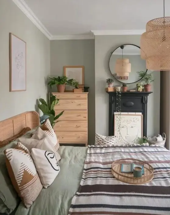 A sage green bedroom with stained furniture, printed bedding, a non working fireplace, a woven pendant lamp and potted greenery