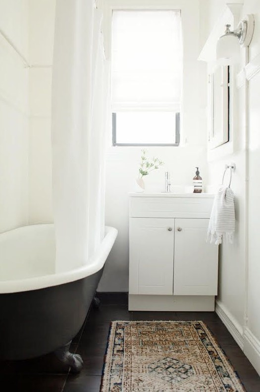 A small boho bathroom with white walls and a dark stained floor, a black clawfoot tub, a white vanity and neutral textiles is a chic idea