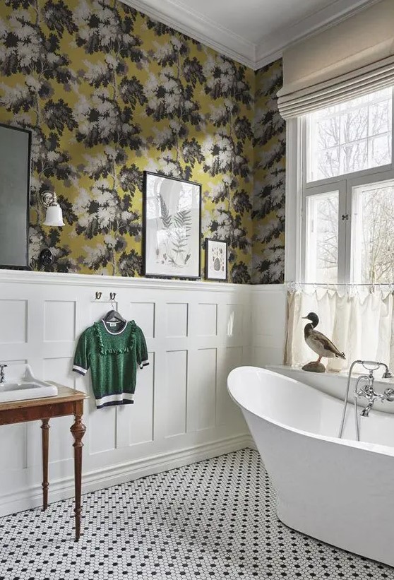 A vintage bathroom with moody floral wallpaper, creamy paneling, a free standing bathtub, black and white tile floor and a vintage vanity