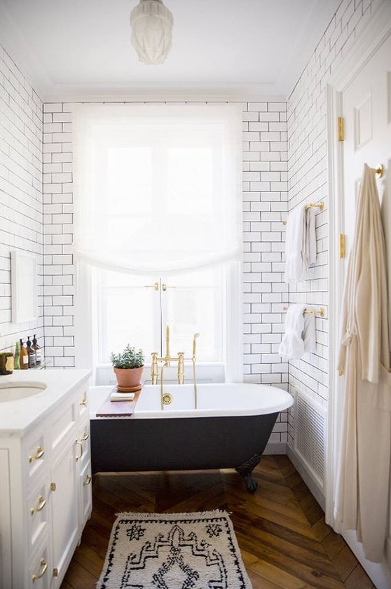 an airy modern bathroom clad with white subway tiles, with a black clawfoot tub, a white vanity with gold touches and gold fixtures