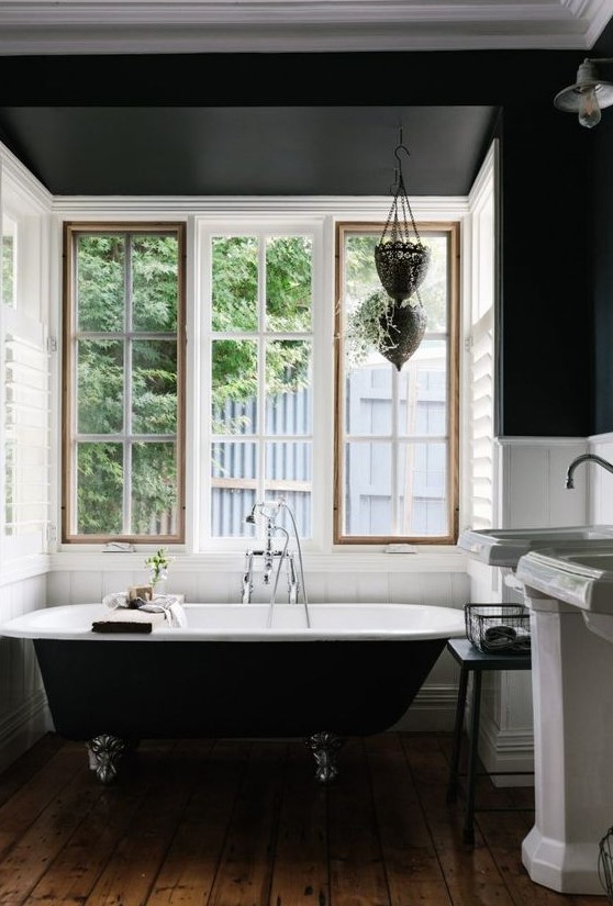 An art deco bathroom with black walls and white planks, a black clawfoot bathtub, free standing sinks and a window